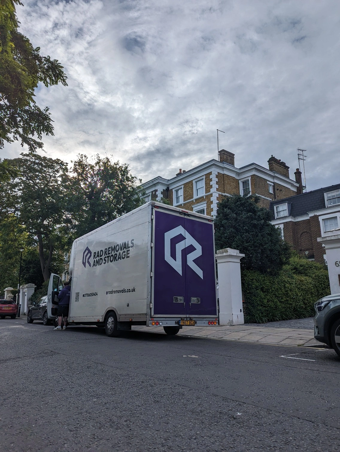 A white RAD van with a bold purple "R" logo parked on a stylish London street, with a sleek modern house featuring large windows and contemporary design in the background under a cloudy London sky, representing time-tested house removals in upscale areas.